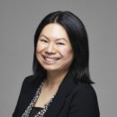 headshot of person smiling, wearing a dark blazer and a patterned blouse, against a plain background.