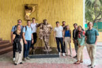 A group of people posing with a bronze filmmaker statue in an outdoor setting with a yellow-striped wall.