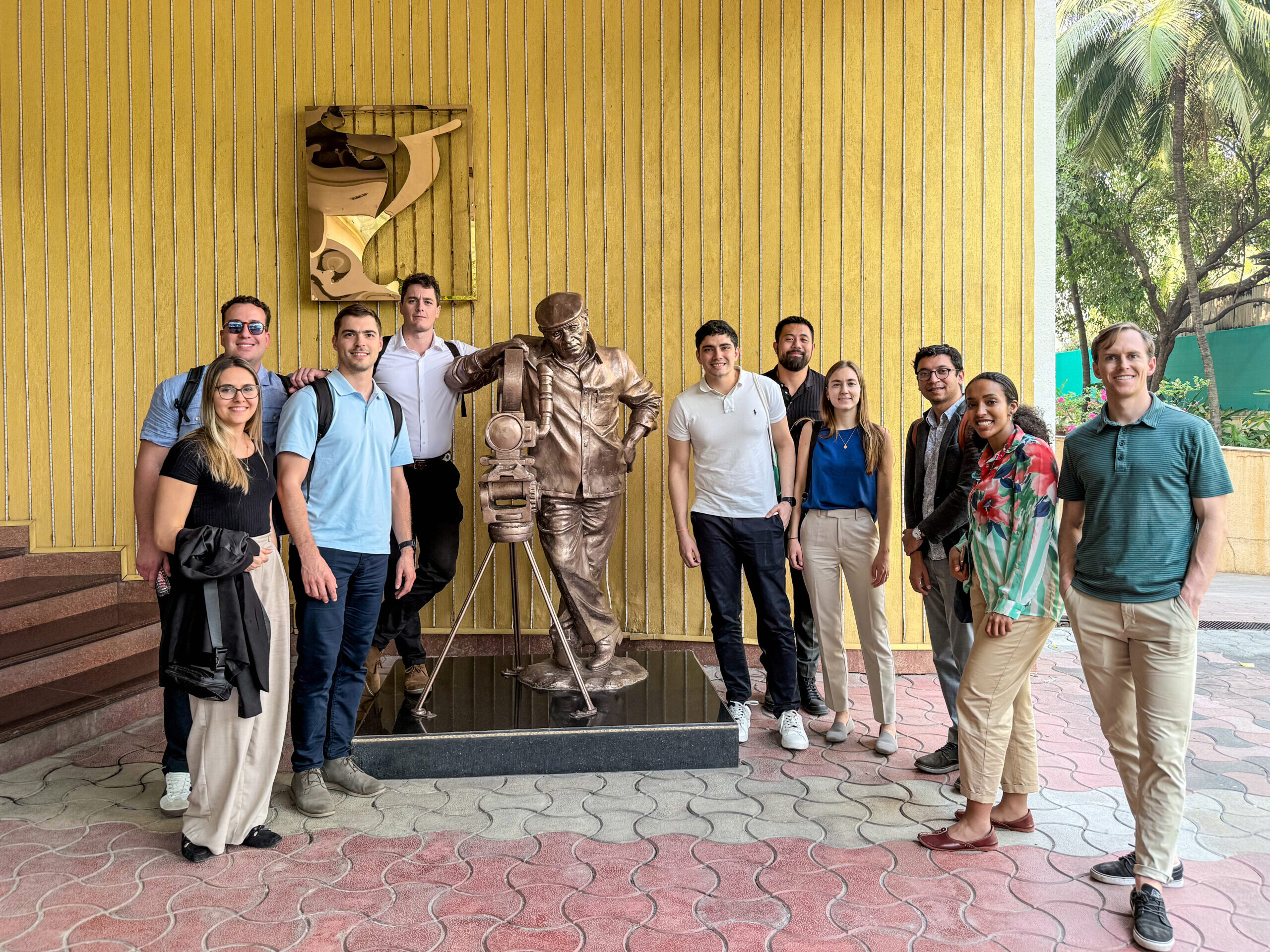 A group of people posing with a bronze filmmaker statue in an outdoor setting with a yellow-striped wall.
